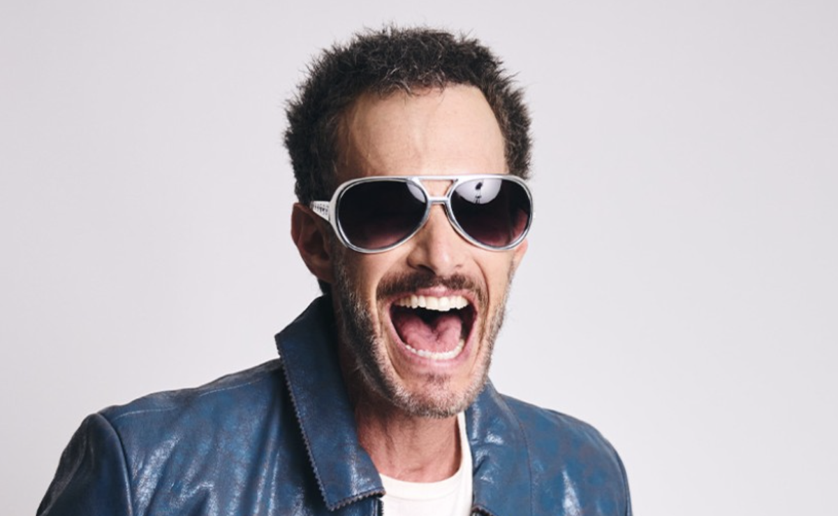 Comedian Josh Wolf laughing exuberantly in sunglasses and a blue jacket against a plain white background in a promo portrait.